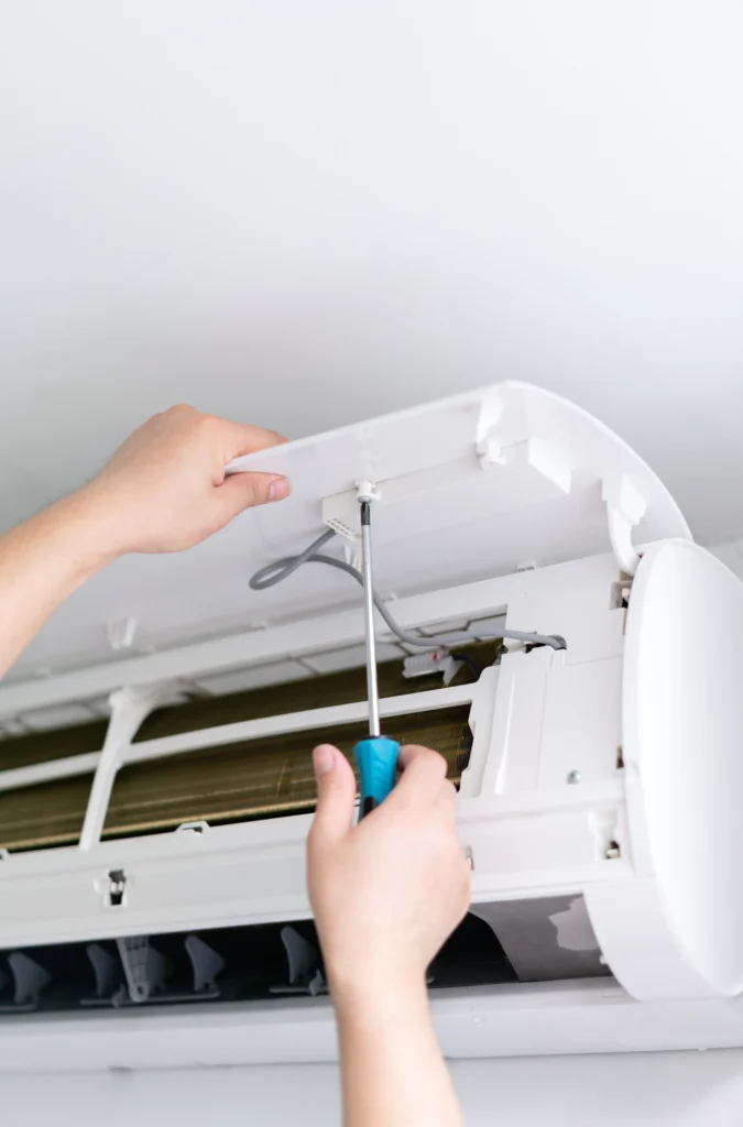 Hands using a screwdriver to repair or install the cover of a wall-mounted air conditioner unit, with exposed internal components visible.